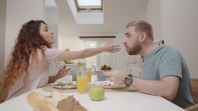 Medium shot of young Caucasian woman using smartphone during dinner with husband at home, annoyed man confiscating it, woman demanding it back and trying to fight, and man throwing it away