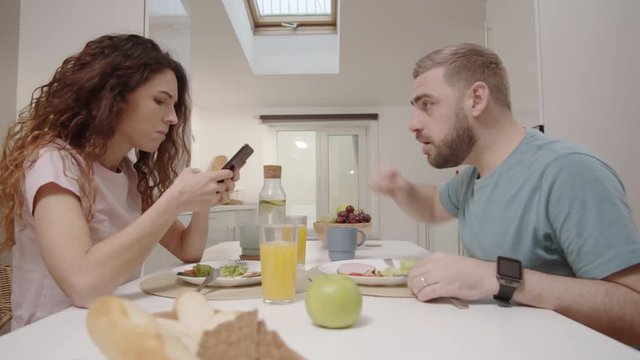 Waist-up shot of distracted Caucasian woman typing messages on smartphone during family meal, jealous husband confiscating phone, checking her private communication and interrogating wife