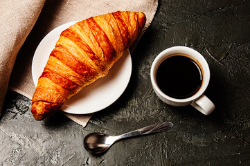 Sweet croissant on saucer, a cup of strong ristretto coffee, a spoon and a linen towel on a dark background