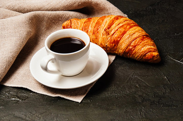 Sweet tasty croissant, white cup of strong ristretto coffee on a saucer and a linen towel on a dark background