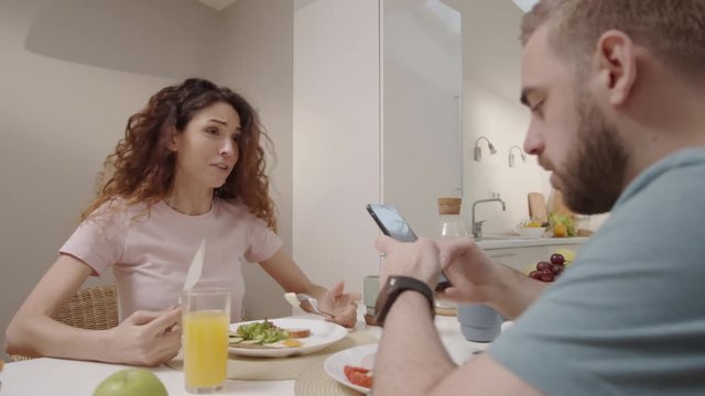 Waist-up arc shot of Caucasian husband using smartphone during family meal, hysterical wife reproaching him, screaming, shouting, grabbing phone and slapping shocked man on face