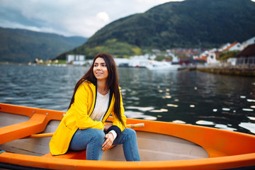 Fototapeta na wymiar The girl tourist in a yellow jacket is sitting in a boat against the backdrop of the mountains in the Norway. Active woman relaxing on the boat by the lake. Travelling, lifestyle, adventure concept.