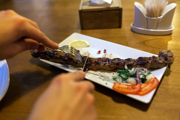 Lula kebab meat dish with vegetables served in restaurant on wooden table. Man cutting and eating barbecue food