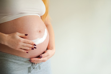 A pregnant woman is applying cream to her stomach for stretch marks. Pregnant tummy, oil for skin elasticity. Pregnant cares for her stomach on a gray background close-up and copy space.