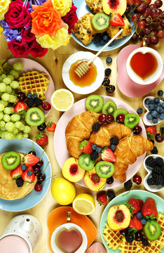 Indulgent breakfast creative layout flat lay with croissants, pancakes, waffles, fruit and roses. Top view vertical.