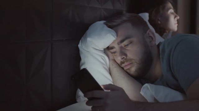 Chest-up panning shot of young Caucasian man lying in bed late at night, holding smartphone with captivated look, and scrolling through news feed, and wife busy with her phone beside