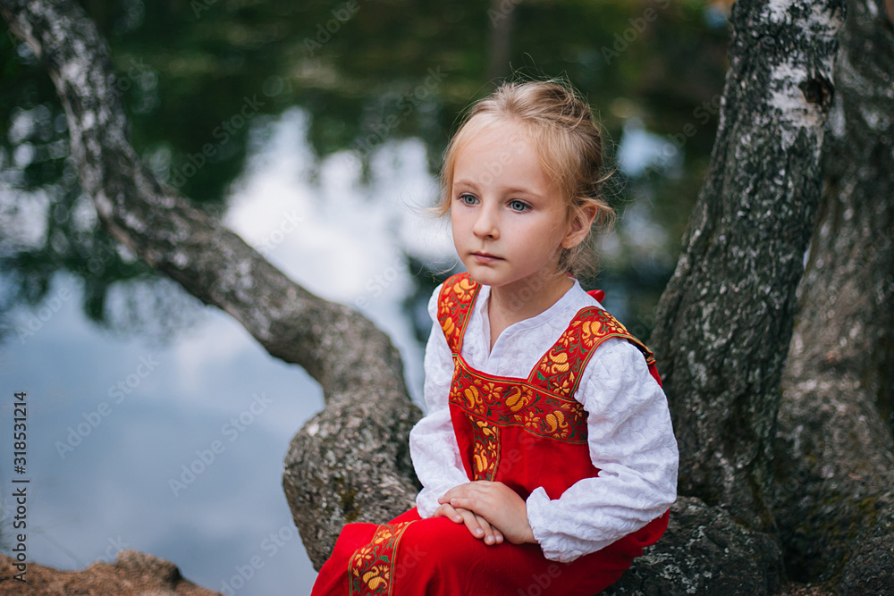 Wall mural Little girl with white hair in a red, Slavic sundress. A girl walks in a birch forest by the pond