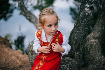 Little girl with white hair in a red, Slavic sundress. A girl walks in a birch forest by the pond