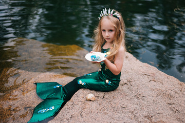 A little girl with white hair with a mermaid tail and shells is sitting on the shore. A little mermaid sits on a large stone by the pond.