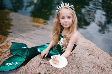 A little girl with white hair with a mermaid tail and shells is sitting on the shore. A little mermaid sits on a large stone by the pond.