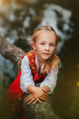 Little girl with white hair in a red, Slavic sundress. A girl walks in a birch forest by the pond