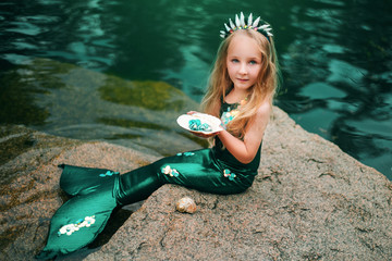 A little girl with white hair with a mermaid tail and shells is sitting on the shore. A little mermaid sits on a large stone by the pond.