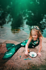 A little girl with white hair with a mermaid tail and shells is sitting on the shore. A little mermaid sits on a large stone by the pond.