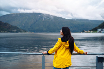 Fototapeta premium The girl tourist in a yellow jacket posing on the lake in Norway. Active woman relaxing near the lake against the backdrop of the mountains in the Norway. Travelling, lifestyle, adventure, wild nature