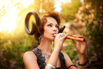 A girl in the image of a faun plays the wooden flute. Model with horns on his head and with furry...