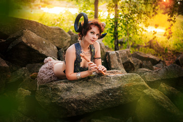 A girl in the image of a faun lies on stones with a wooden flute. Model with horns on the head and with furry pants in the forest - obrazy, fototapety, plakaty