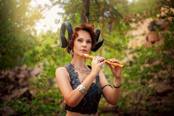 A girl in the image of a faun stands with a wooden flute. Model with horns on his head and with furry pants in the woods among the stones - obrazy, fototapety, plakaty