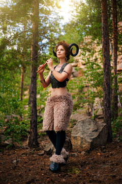 A girl in the image of a faun stands with a wooden flute. Model with horns on his head and with furry pants in the woods among the stones
