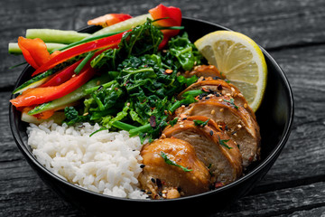 Buddha bowl meal with teriyaki chicken fillet, fresh vegetable salad, stew cabbage and basmati rice on a rustic table. Delicious dish in a black bowl, close-up shot.