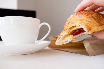 Female hand holding freshly bitten croissant, next to cup of coffee on laptop. On a white background. Concept of learning