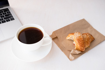 Layout of white cup of coffee, bitten croissant near laptop on a white table.top view