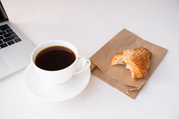 Layout of white cup of coffee, bitten croissant near laptop on a white table.top view