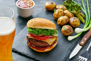 Delicious cheeseburger with baked potatoes and broccoli on a black stone board served with a glass of lager beer.