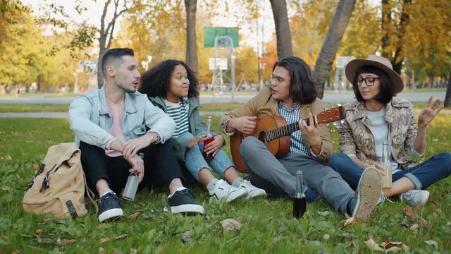 Young men and women friends are playing the guitar singing enjoying warm autumn day in urban park. Modern lifestyle, music and happiness concept.