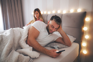 Couple lie in bed and man using phone.