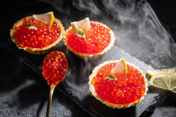 Tartlets with red caviar served on a black background. Delicious seafood. Steam and hard light