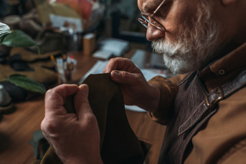 attentive, senior shoemaker holding piece of genuine leather in workshop