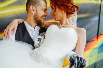 Groom holding bride on his hands in front of graffiti wall