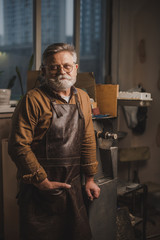 positive, bearded shoemaker in leather apron looking at camera while standing in workshop