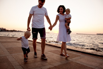 Fototapeta premium happy family are walking with toddler twins on the city promenade against the sea and sunset.