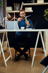 Young innovative caucasian bearded engineer sitting at his modern office and holding windmill model. Sustainable concept.