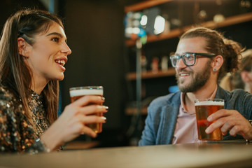 Young handsome caucasian bearded man with eyeglasses leaning on bar counter in pub, drinking beer and chatting with friend.