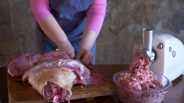 beautiful mature woman in pink sweater and gray economic apron cuts large piece of pork into pieces with knife and ax. girl puts small pieces in meat grinder and grinds them into minced meat