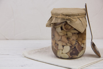 canned mushrooms in a jar of garlic black pepper, a spoon stands on a white cotton napkin on a white wooden background