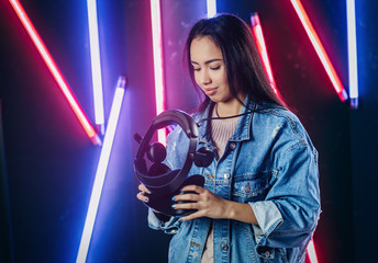 Beautiful young girl holds in hands virtual reality glasses in the dark studio with neon light 
