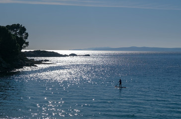 The man on the SUP board is floating in the bay in blue sea at sunny day. Active recreation in nature. A man spends a day enjoying SUP ride on the sea. Active lifestyle. Amazing bays of Catalonia.