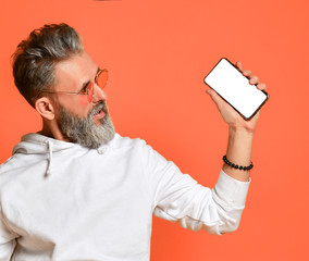 Stylish smiling grey-haired hipster adult man in white hoodie and sunglasses standing looking at mobile cell phone smartphone in hand with blank screen for text space