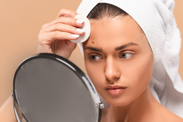 young woman holding cotton pad while looking at mirror isolated on beige