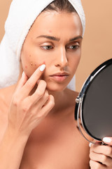  nude girl in towel touching face with pimples and looking at mirror isolated on beige