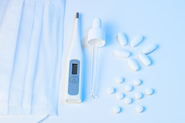 electronic thermometer, protective mask, pipette and tablets lie on white background with blue illumination, epidemic