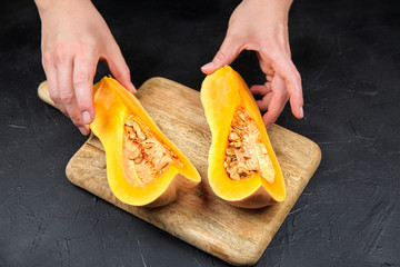 Pumpkin in female hands, butternut squash pieces with seeds on wooden cutting board on black...