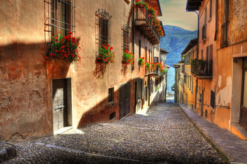 Narrow Street with Sunlight and Old Houses with Red Flowers and Lake with Mountain in Cannobio,...
