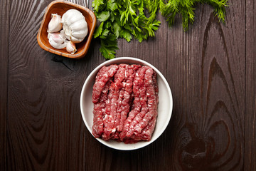 Cooking minced meat with fresh seasonings on a wooden table, flat lay. White garlic heads and cloves, green dill and parsley leaves. Bowl with raw ground beef and condiments, top view
