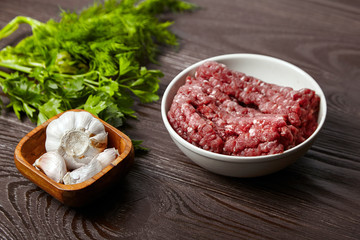 Cooking minced meat with fresh seasonings on a wooden table. White garlic heads and cloves, green dill and parsley leaves. Bowl with raw ground beef and condiments on brown wood background
