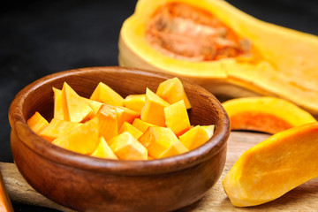 Butternut squash pieces, wooden bowl with pumpkin seeds, cutting board on black background, closeup. Cooking winter squash