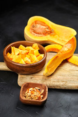 Butternut squash pieces, wooden bowl with pumpkin seeds, cutting board on black background, closeup. Cooking winter squash